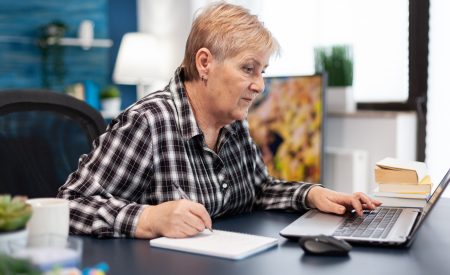 Mature entrepreneur taking notes on notebook working in home office. Elderly woman in home living room using moder technoloy laptop for communication sitting at desk indoors.
